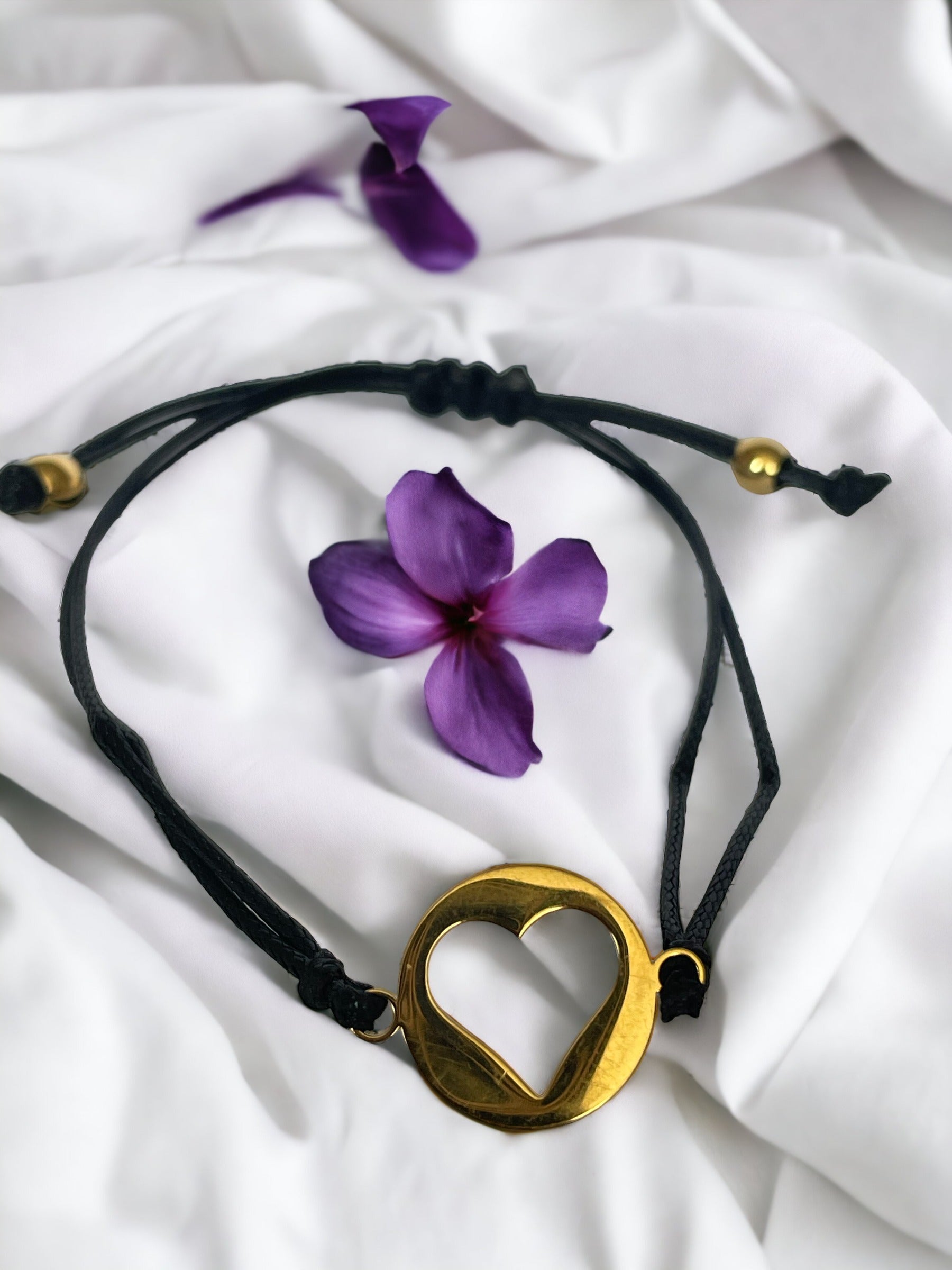 This bracelet features a black cord made of high-quality material, complemented by a stainless steel pendant. The pendant has a circular shape with a heart shape in, plated in gold, adding a touch of elegance and sentiment to your style."