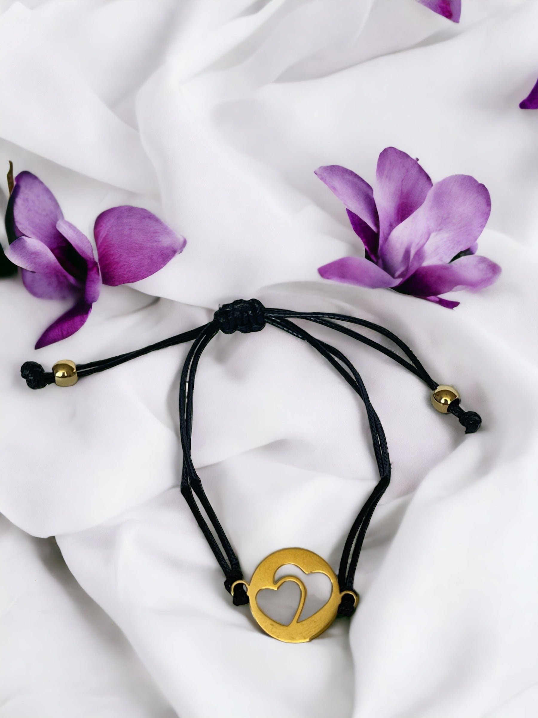 This bracelet features a black cord made of high-quality material, complemented by a stainless steel pendant. The pendant has a circular shape with two  heart shape in, plated in gold, adding a touch of elegance and sentiment to your style."