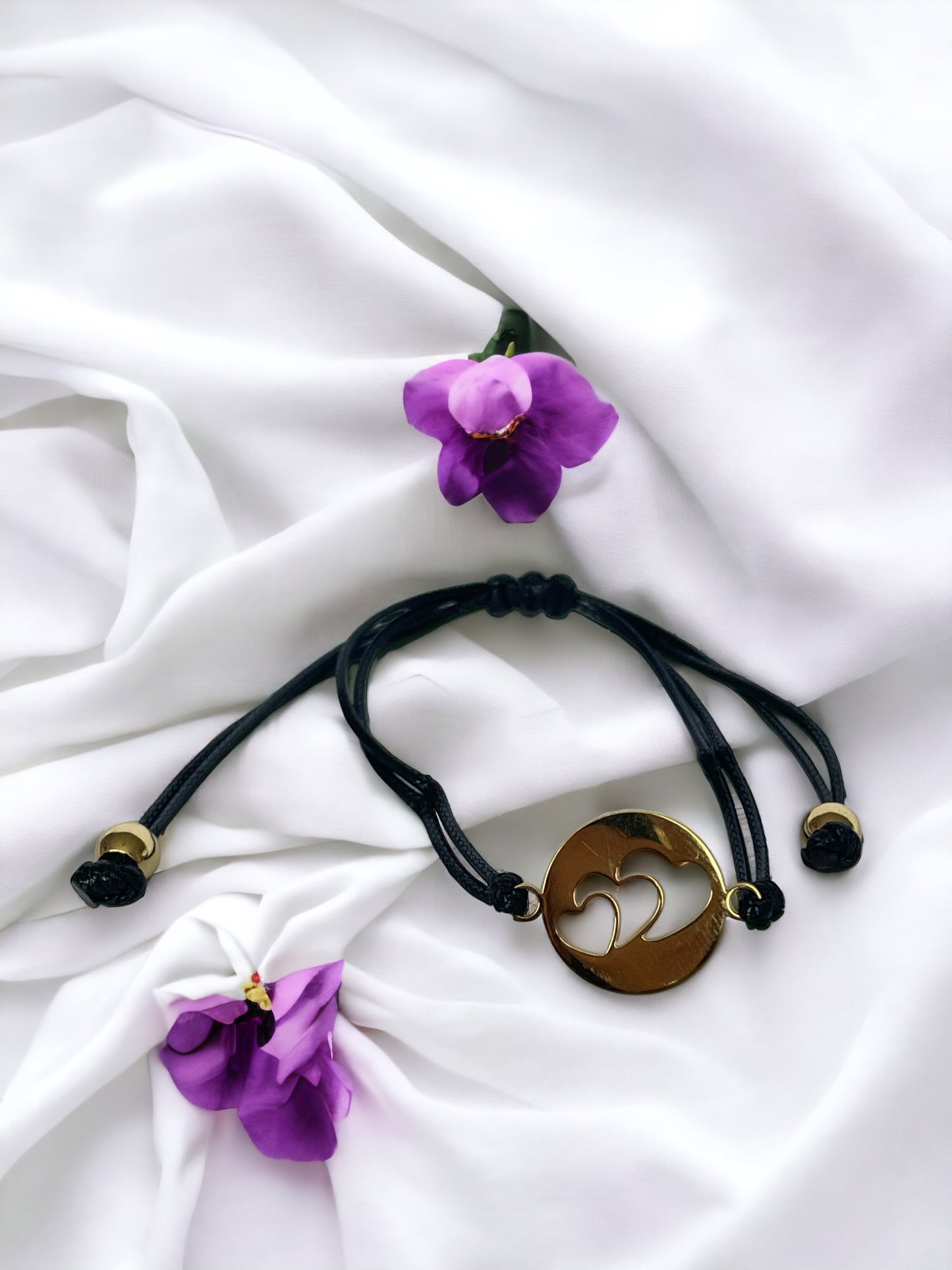 This bracelet features a black cord made of high-quality material, complemented by a stainless steel pendant. The pendant has a circular shape with three heart shape in, plated in gold, adding a touch of elegance and sentiment to your style