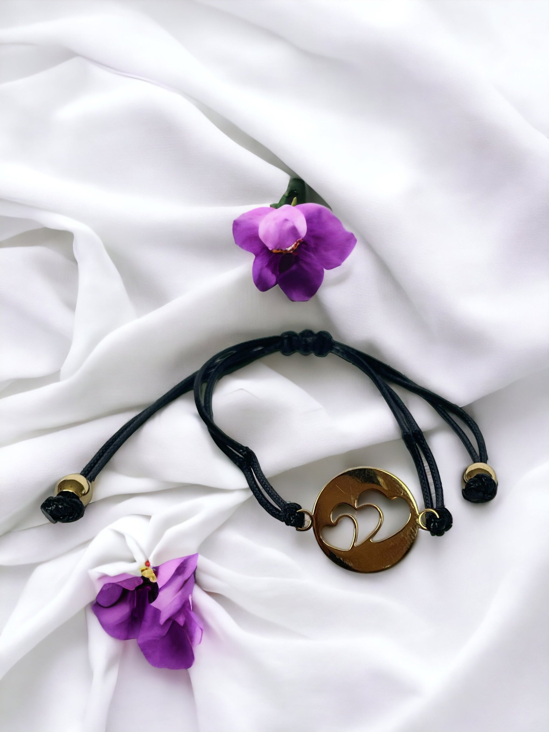 This bracelet features a black cord made of high-quality material, complemented by a stainless steel pendant. The pendant has a circular shape with three heart shape in, plated in gold, adding a touch of elegance and sentiment to your style