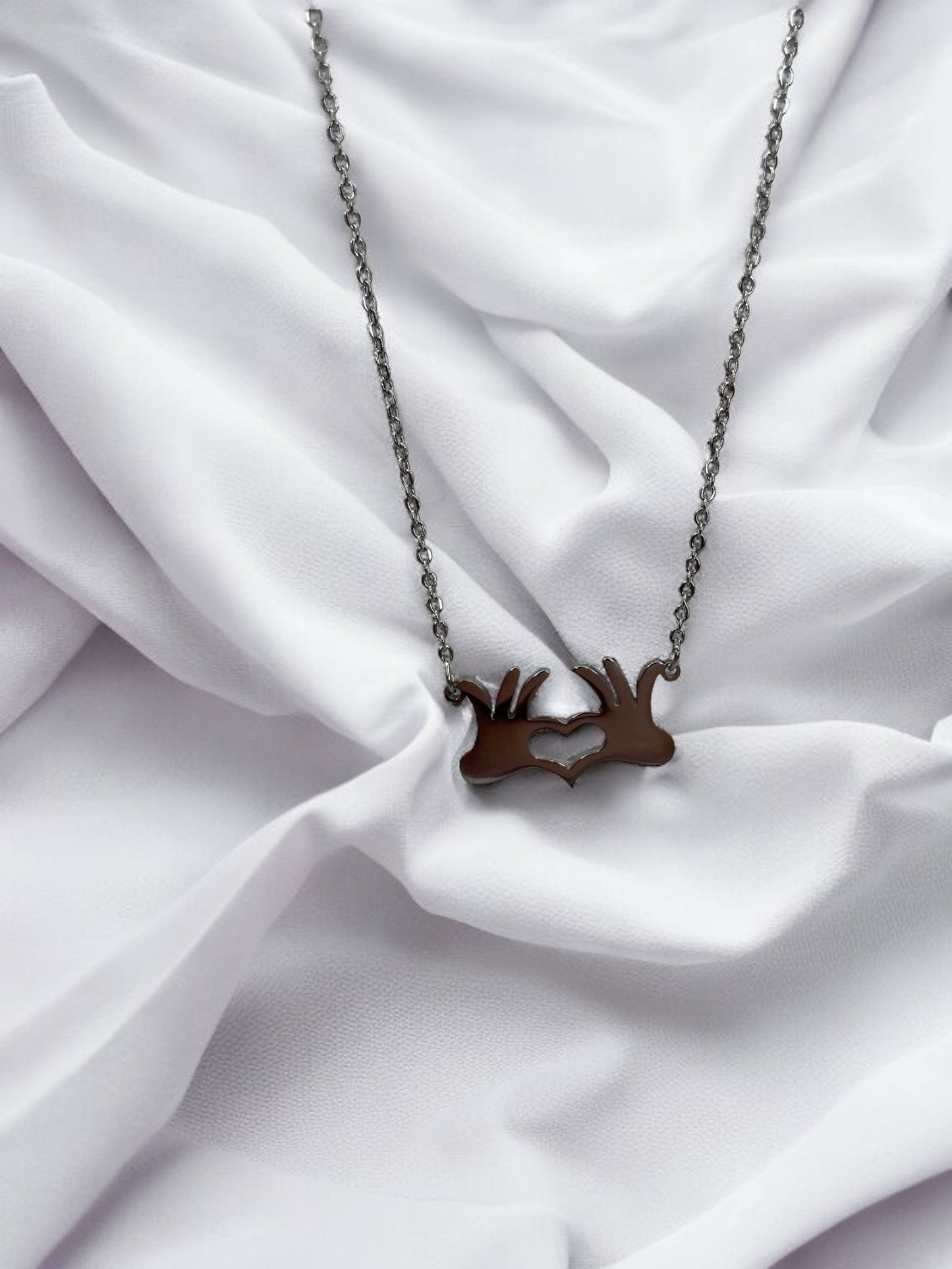 This is a visual description of a pendant necklace. The necklace features a unique pendant that illustrates two hands joined together at the wrists, creating a heart shape in the open space between them. The necklace is available in either a shiny stainless steel or a gold-plated finish, providing options for personal preference. The linked chain complements the pendant's message of connection and heartfelt togetherness.