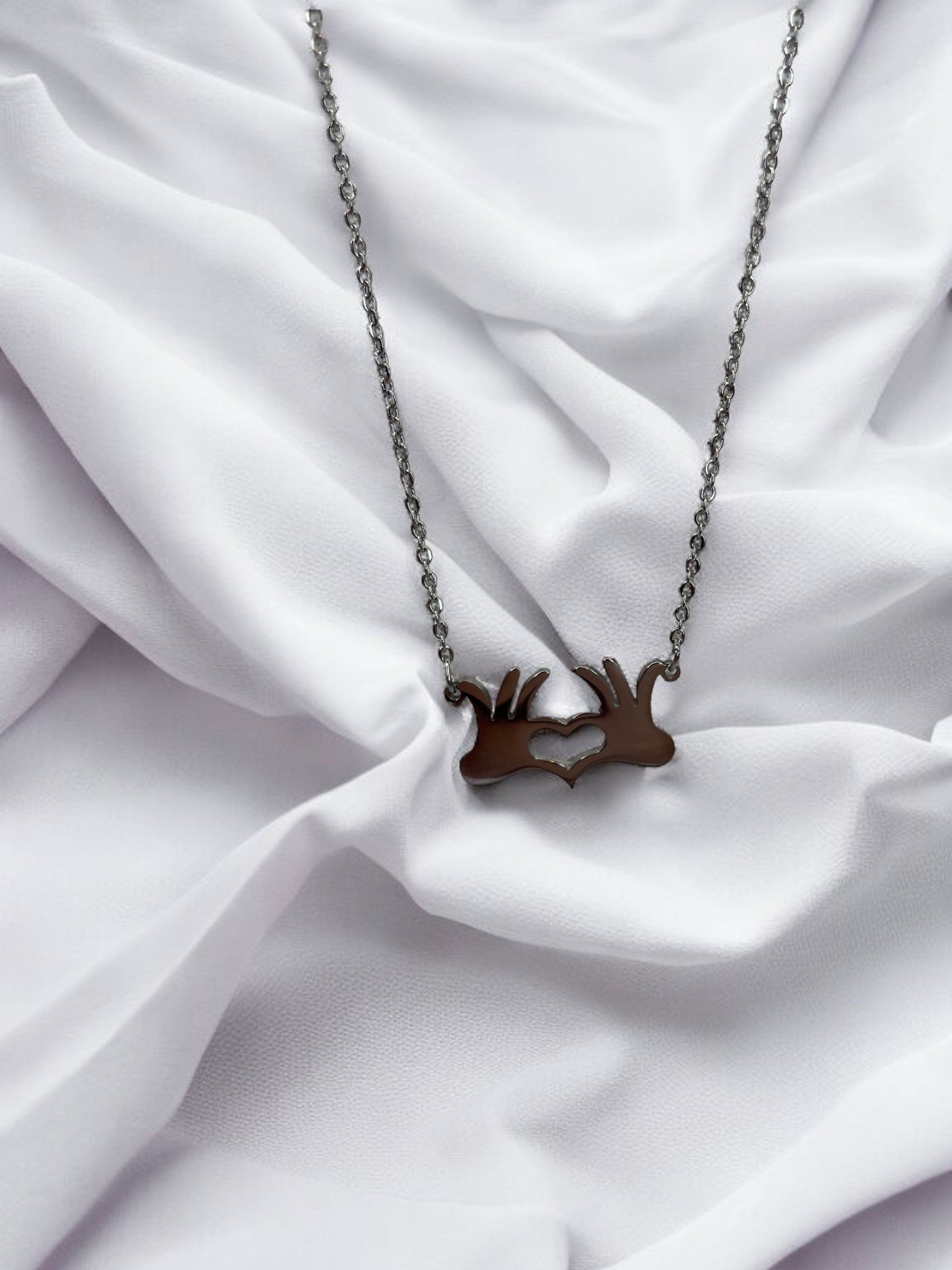 This is a visual description of a pendant necklace. The necklace features a unique pendant that illustrates two hands joined together at the wrists, creating a heart shape in the open space between them. The necklace is available in either a shiny stainless steel or a gold-plated finish, providing options for personal preference. The linked chain complements the pendant's message of connection and heartfelt togetherness.