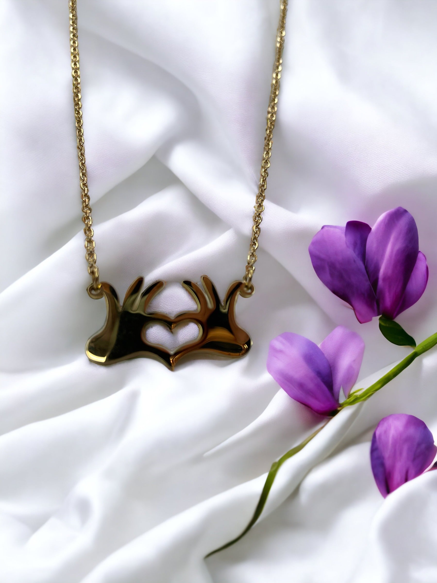This is a visual description of a pendant necklace. The necklace features a unique pendant that illustrates two hands joined together at the wrists, creating a heart shape in the open space between them. The necklace is available in either a shiny stainless steel or a gold-plated finish, providing options for personal preference. The linked chain complements the pendant's message of connection and heartfelt togetherness.