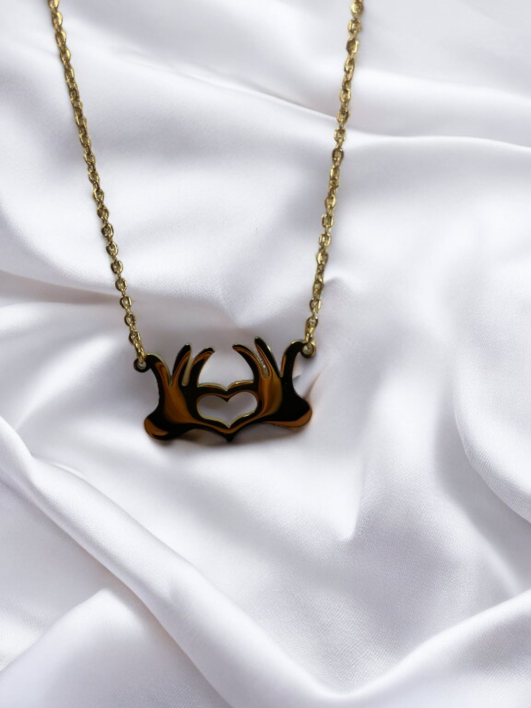 This is a visual description of a pendant necklace. The necklace features a unique pendant that illustrates two hands joined together at the wrists, creating a heart shape in the open space between them. The necklace is available in either a shiny stainless steel or a gold-plated finish, providing options for personal preference. The linked chain complements the pendant's message of connection and heartfelt togetherness.
