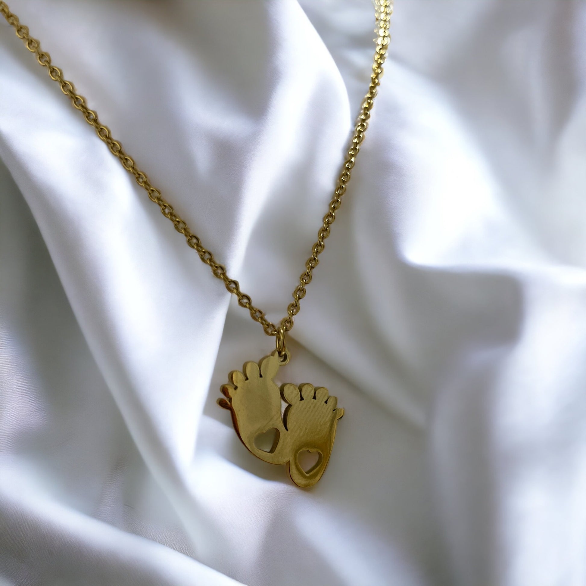 This image shows a necklace with two footprint-shaped pendants linked together, each cutout with a heart, expressing the purest form of love. Cast in a golden hue, the pendants dangle from a delicate gold-tone chain. They are also available in a silver-tone stainless steel option, giving a choice of styles for the wearer. This piece is a tender celebration of growth and affection, making it a heartfelt accessory or gift.