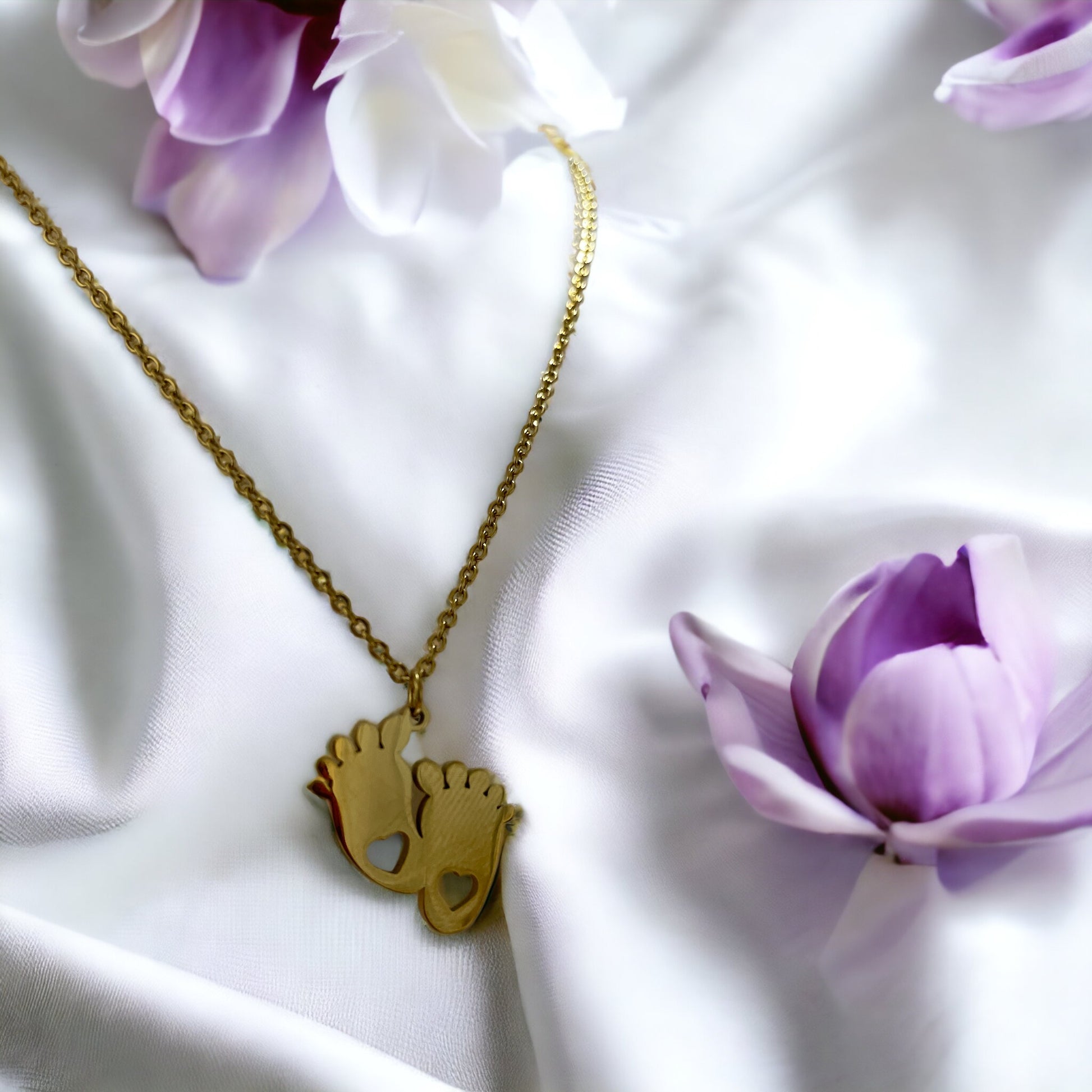 This image shows a necklace with two footprint-shaped pendants linked together, each cutout with a heart, expressing the purest form of love. Cast in a golden hue, the pendants dangle from a delicate gold-tone chain. They are also available in a silver-tone stainless steel option, giving a choice of styles for the wearer. This piece is a tender celebration of growth and affection, making it a heartfelt accessory or gift.