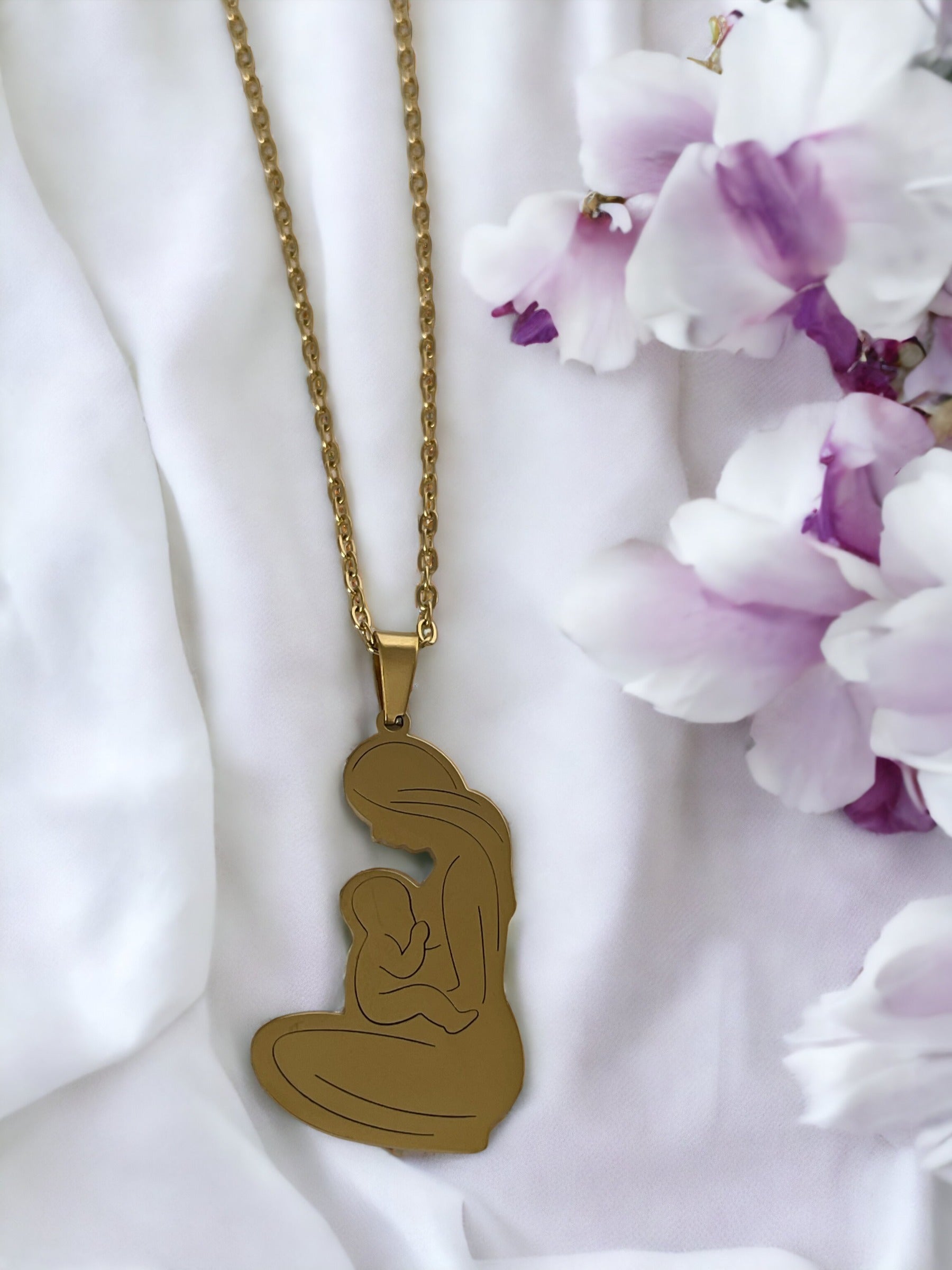 This image displays the Maternal Grace Pendant, available in both a reflective stainless steel and a warm gold-plated finish. The pendant is heart-shaped, featuring a stylized outline of a mother embracing her infant. It dangles from a complementary chain, set against a backdrop of soft white fabric interspersed with delicate purple blooms, highlighting the pendant's beauty and the sentiment it represents.