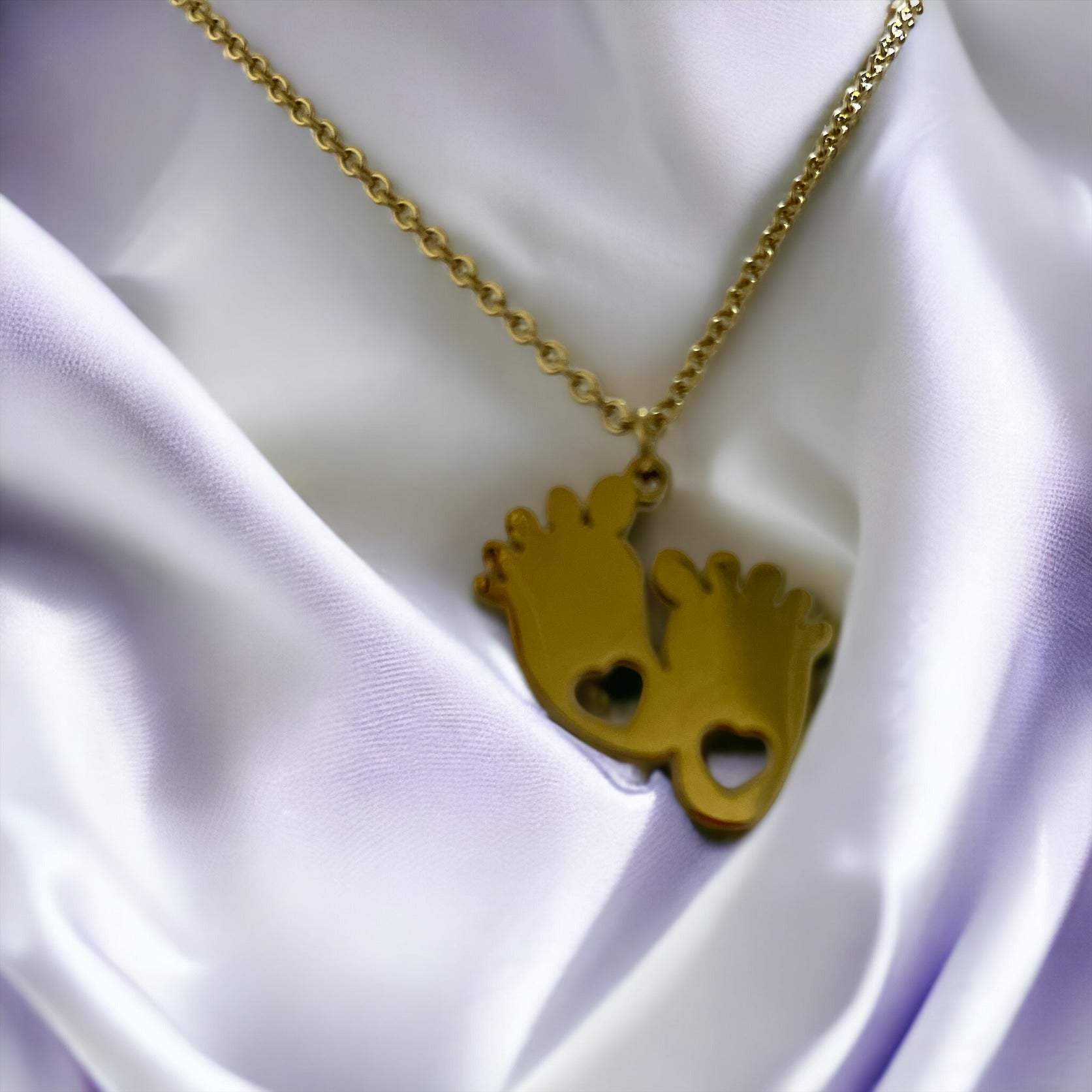 This image shows a necklace with two footprint-shaped pendants linked together, each cutout with a heart, expressing the purest form of love. Cast in a golden hue, the pendants dangle from a delicate gold-tone chain. They are also available in a silver-tone stainless steel option, giving a choice of styles for the wearer. This piece is a tender celebration of growth and affection, making it a heartfelt accessory or gift.