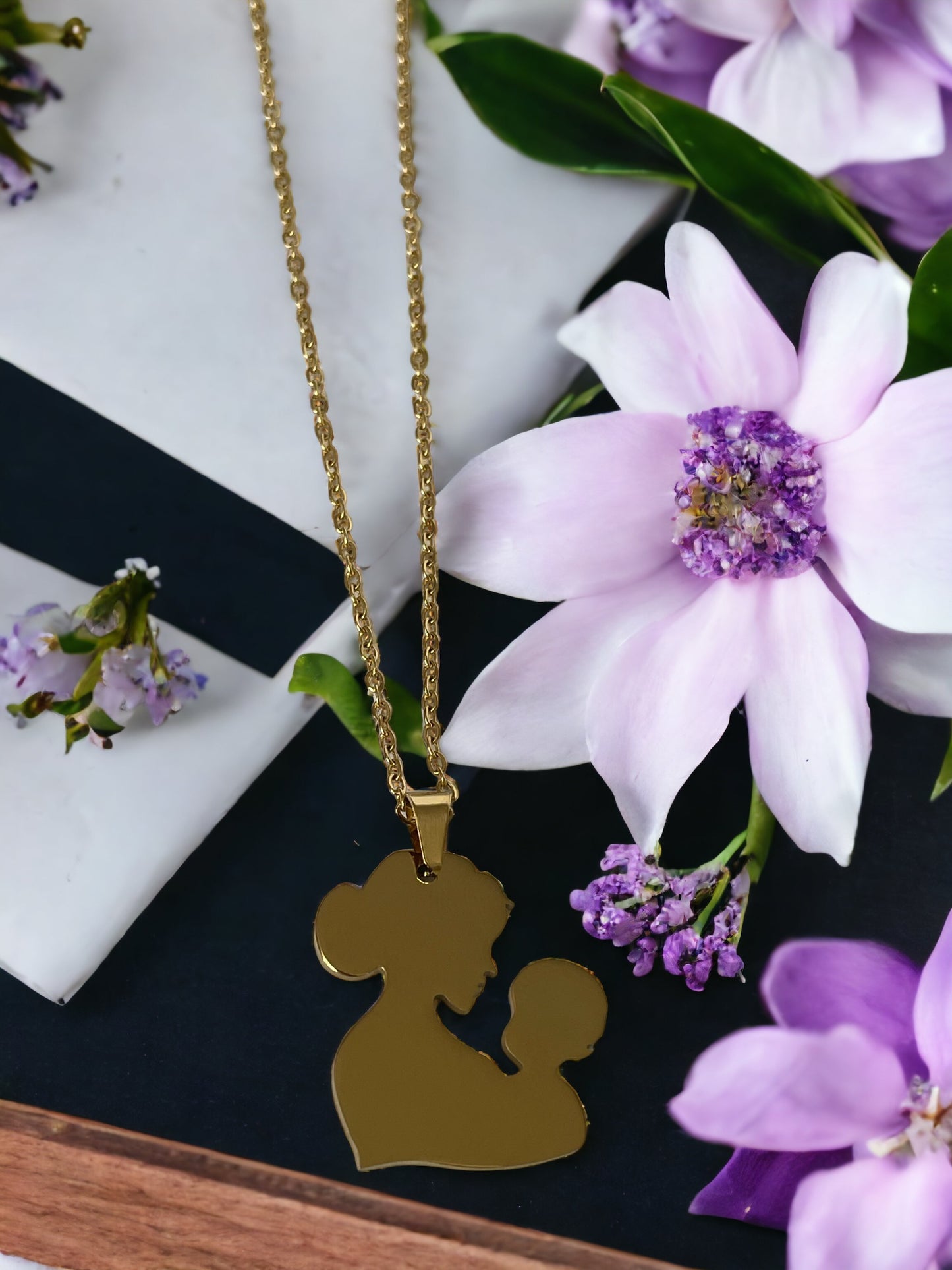 You are viewing a necklace with a pendant that artistically represents a mother holding her child. The pendant's design is abstract, with smooth, flowing lines that outline the figures, merging into a single form. The pendant is golden in color, hinting at a gold-plated material, although it is also available in stainless steel. It hangs on a matching golden chain that complements the pendant's warm tones. This piece is designed to embody the warmth and love of the maternal bond.
