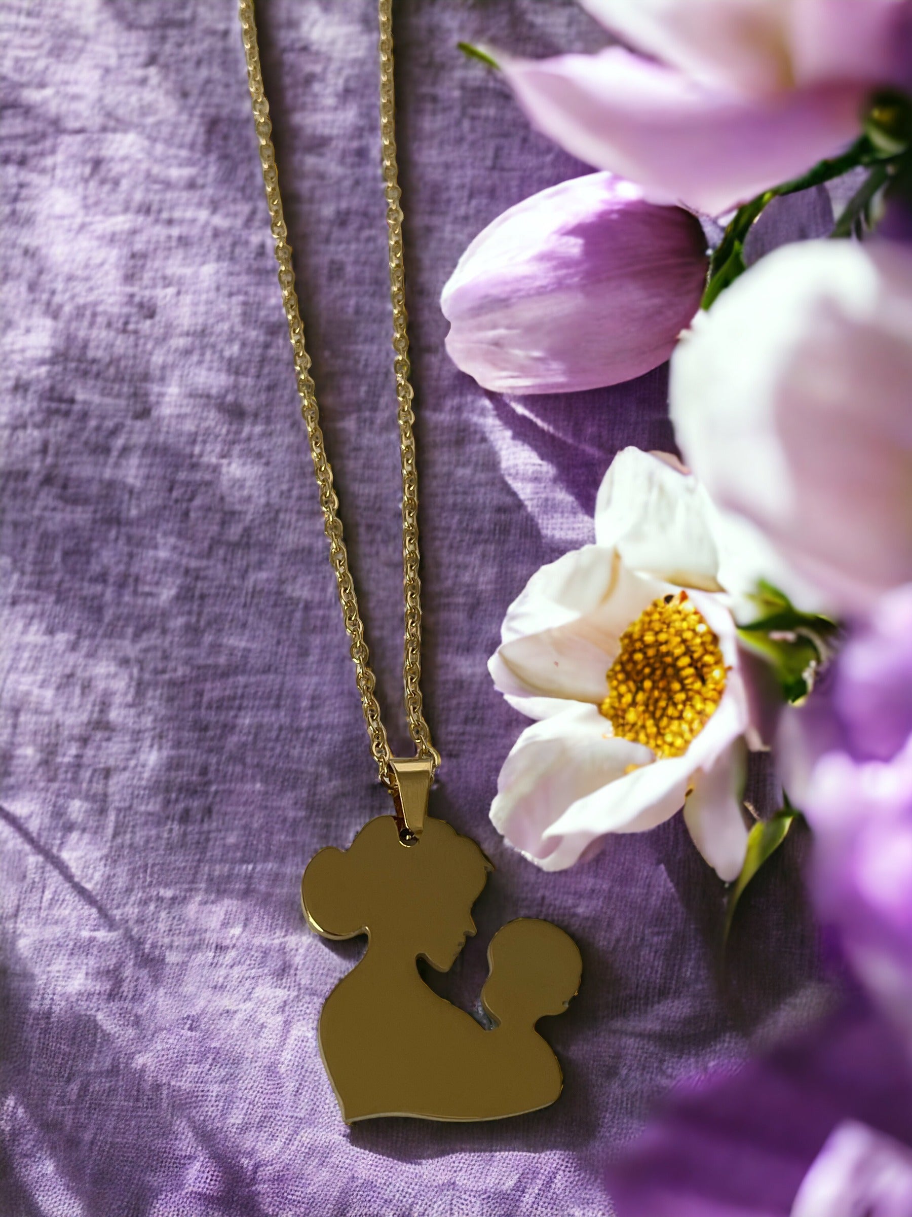 You are viewing a necklace with a pendant that artistically represents a mother holding her child. The pendant's design is abstract, with smooth, flowing lines that outline the figures, merging into a single form. The pendant is golden in color, hinting at a gold-plated material, although it is also available in stainless steel. It hangs on a matching golden chain that complements the pendant's warm tones. This piece is designed to embody the warmth and love of the maternal bond.
