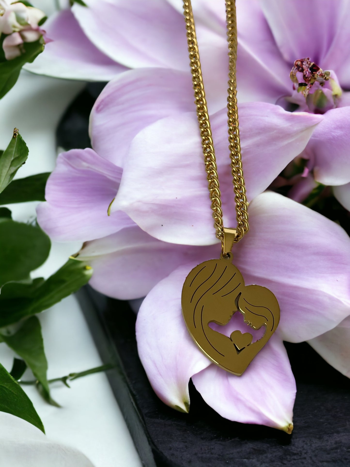 This image features the "Maternal Embrace Heart Pendant," a beautifully crafted piece with a silhouette of a mother and her child in a tender embrace, forming a heart shape. The pendant has a rich, golden hue, suggesting it is available in gold plating, with an option for stainless steel as well. It is draped over a delicate, magnolia petal, surrounded by soft, pastel flowers, enhancing the pendant's elegance and the sentiment it represents.