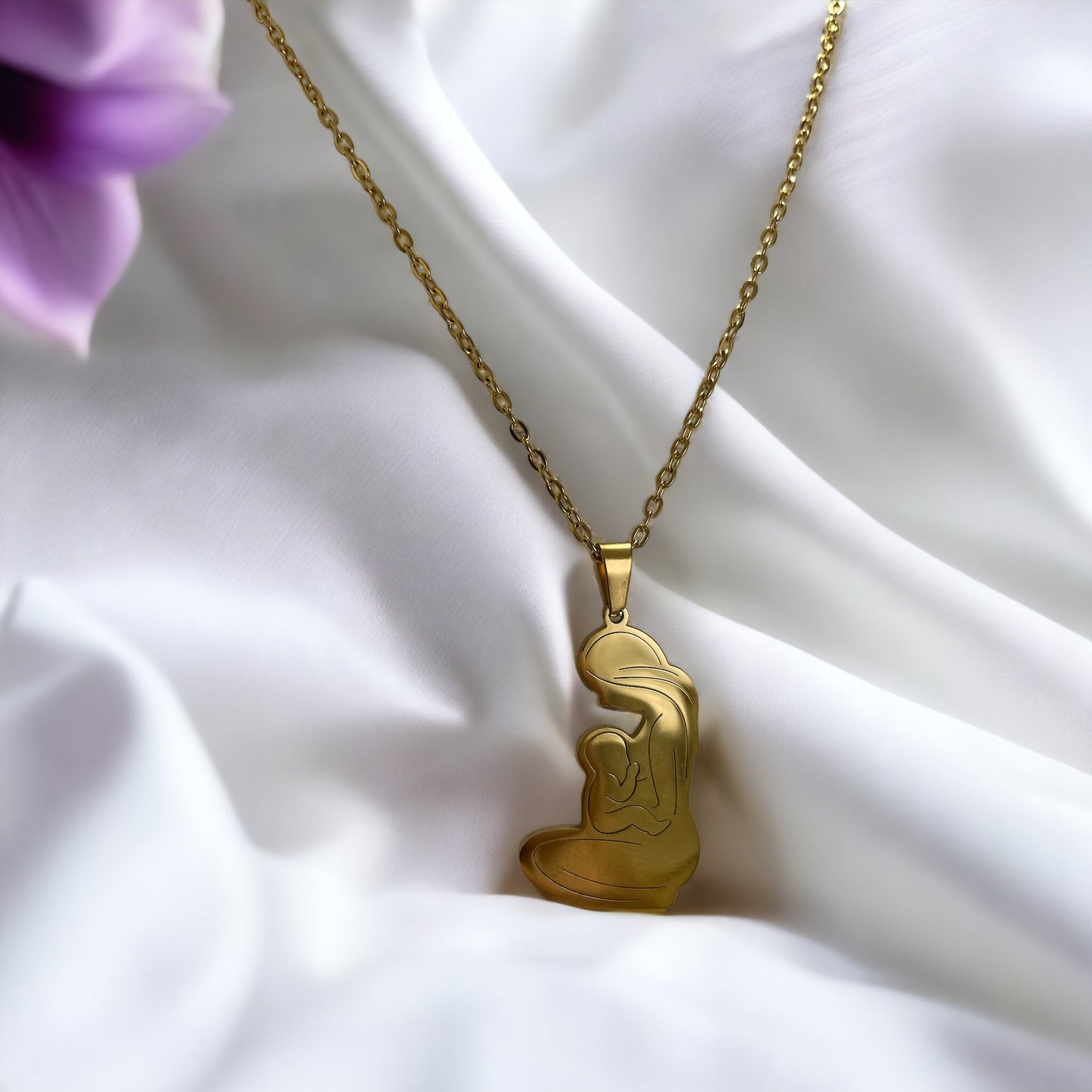 This image displays the Maternal Grace Pendant, available in both a reflective stainless steel and a warm gold-plated finish. The pendant is heart-shaped, featuring a stylized outline of a mother embracing her infant. It dangles from a complementary chain, set against a backdrop of soft white fabric interspersed with delicate purple blooms, highlighting the pendant's beauty and the sentiment it represents.