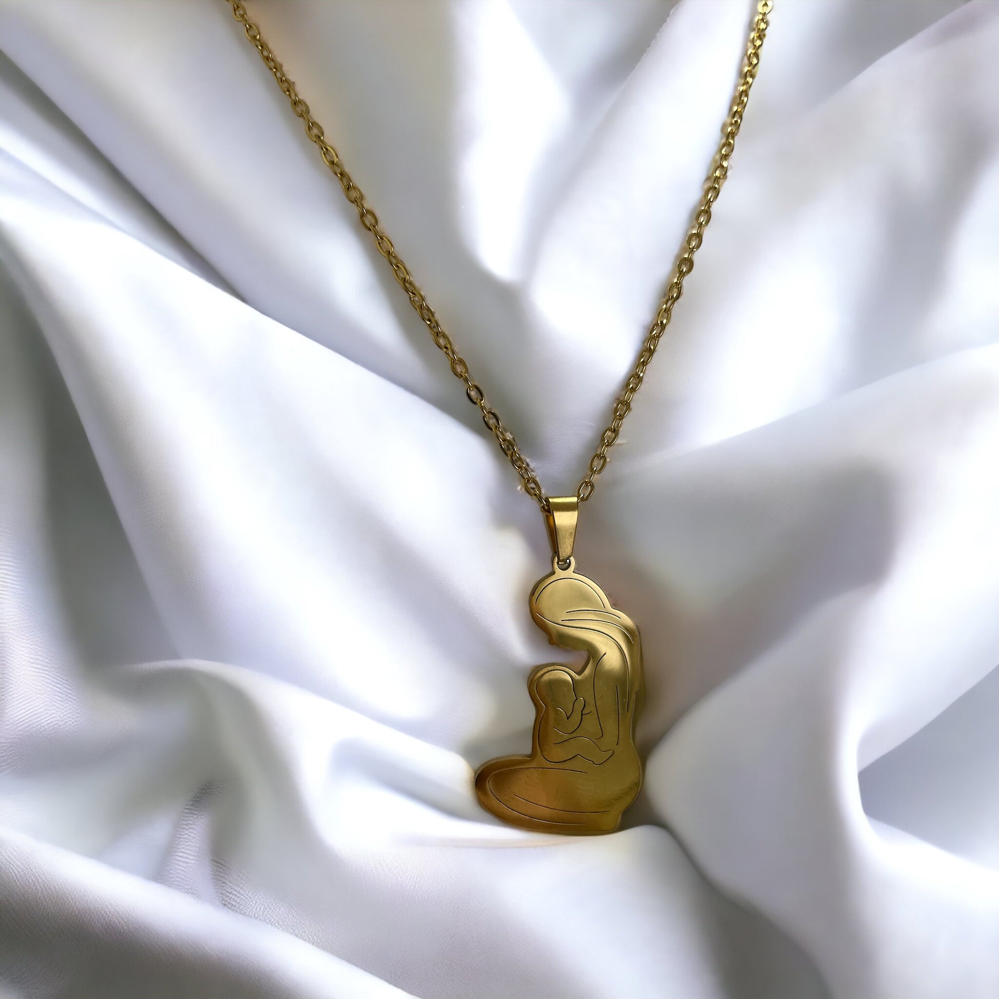 This image displays the Maternal Grace Pendant, available in both a reflective stainless steel and a warm gold-plated finish. The pendant is heart-shaped, featuring a stylized outline of a mother embracing her infant. It dangles from a complementary chain, set against a backdrop of soft white fabric interspersed with delicate purple blooms, highlighting the pendant's beauty and the sentiment it represents.