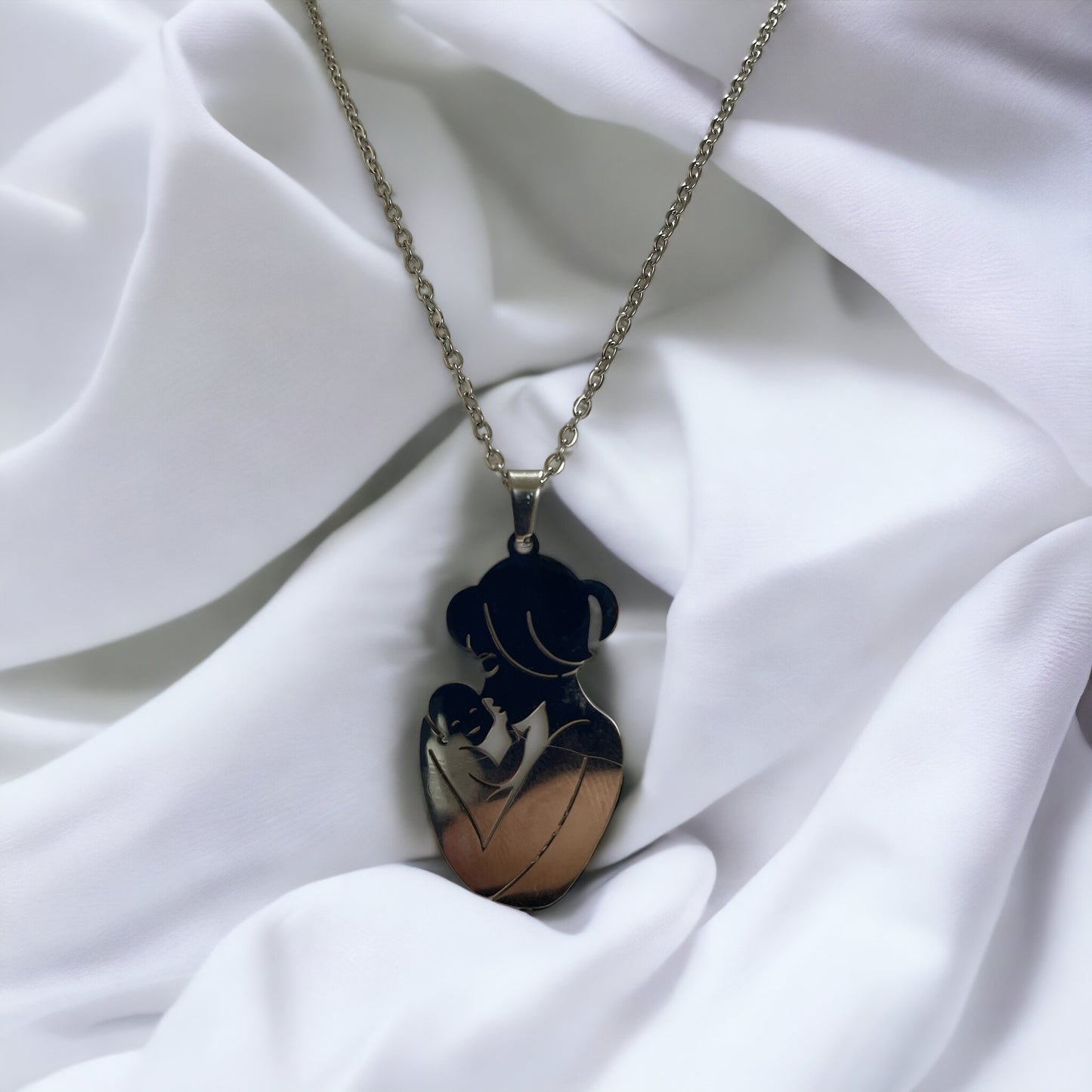  The image displays a necklace with a pendant featuring the silhouette of a mother holding her baby close. The design is simple yet evocative, capturing a moment of tenderness between parent and child. The pendant is available in two finishes: a lustrous stainless steel and a warm gold plating. It hangs from a matching chain, complementing the pendant's finish. The necklace embodies a sense of nurturing love and would make a thoughtful gift or a personal keepsake, symbolizing the bond of motherhood.