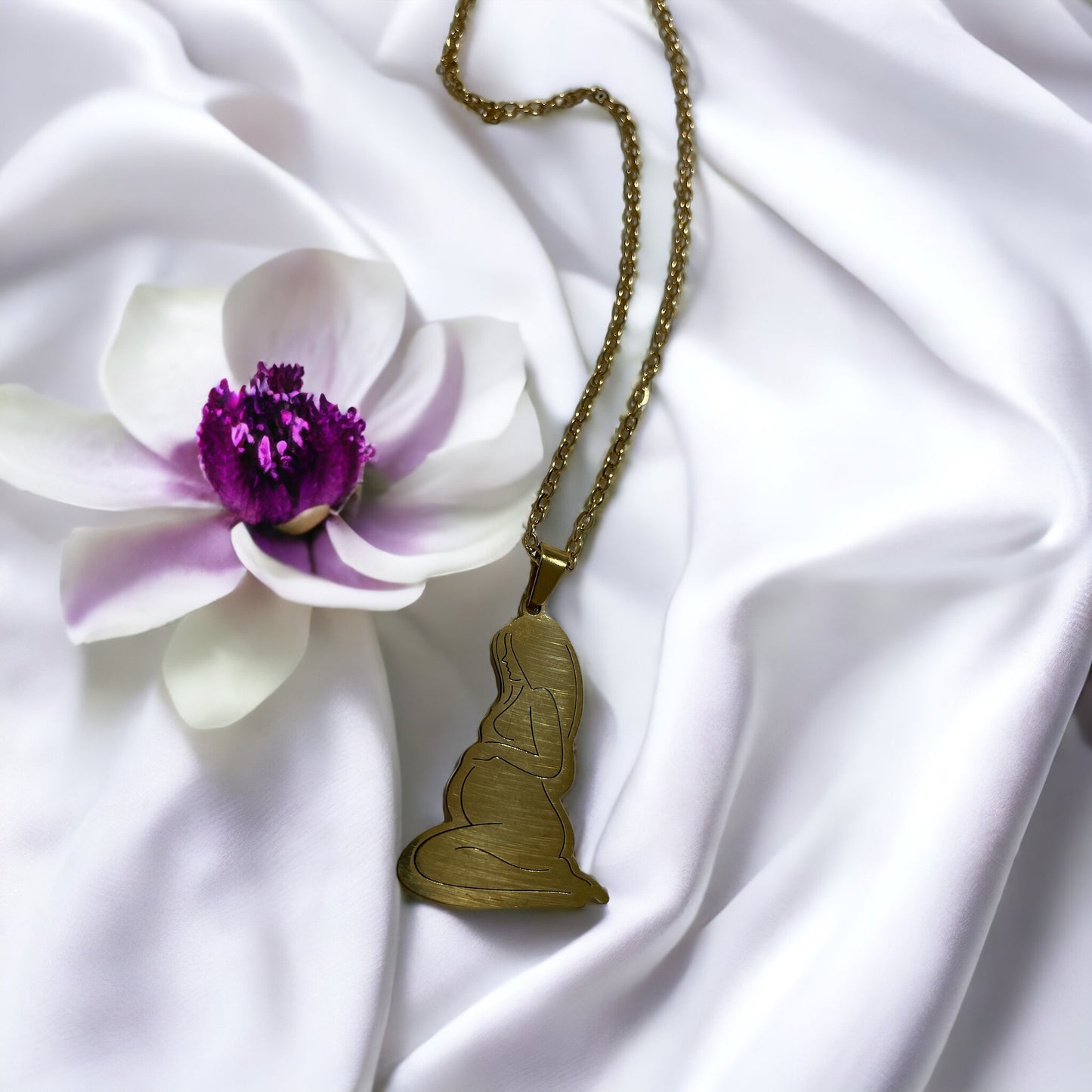 This image presents the Serene Motherhood Pendant, a delicate and polished pendant in gold plating that can also be chosen in stainless steel. The pendant features the graceful outline of a mother in a contemplative pose, cradling her child in a tender embrace, symbolizing the protective and loving bond shared. It hangs from a matching chain, showcased against a backdrop of soft, white satin that gently drapes around it, accentuating the pendant's smooth contours and reflective surface.
