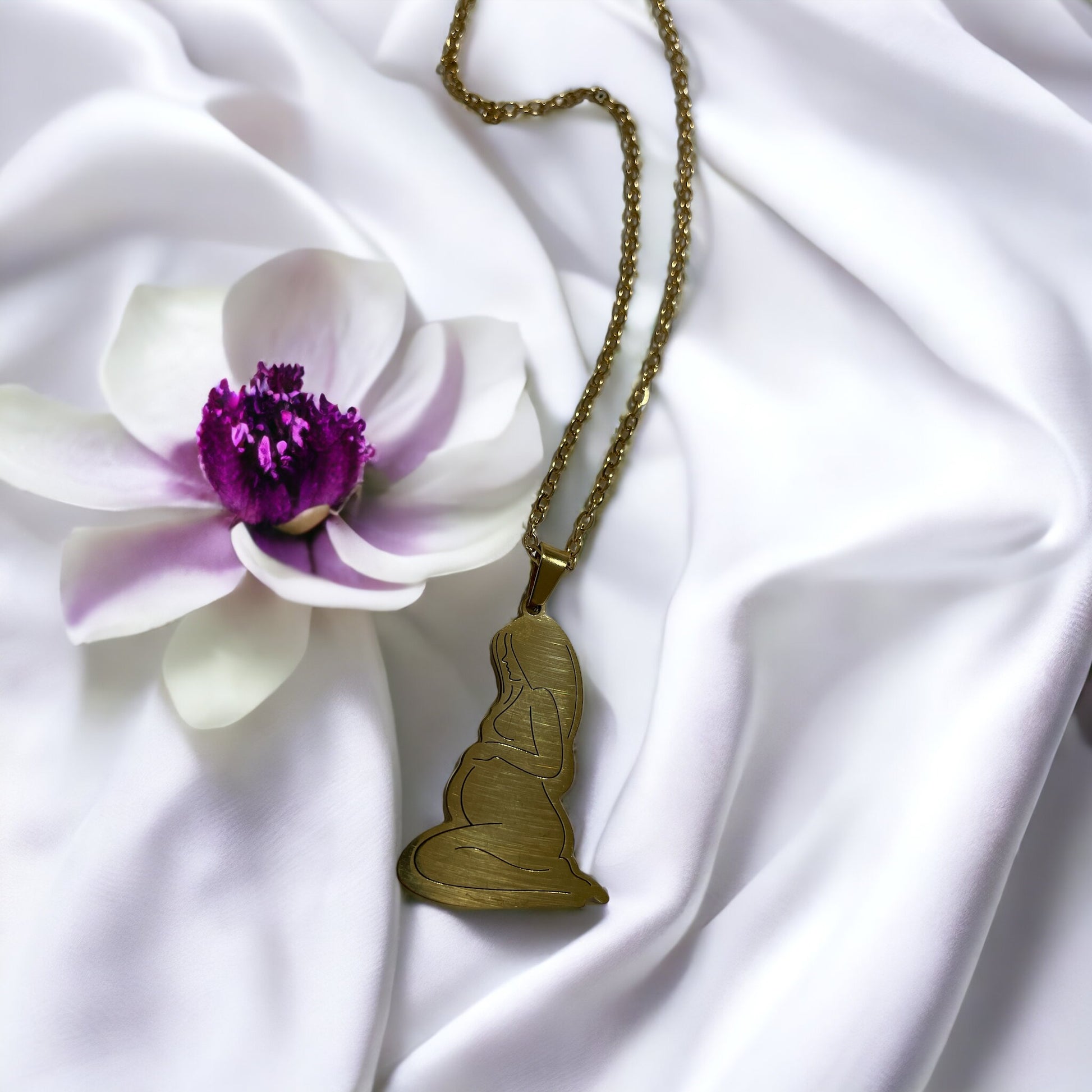 This image presents the Serene Motherhood Pendant, a delicate and polished pendant in gold plating that can also be chosen in stainless steel. The pendant features the graceful outline of a mother in a contemplative pose, cradling her child in a tender embrace, symbolizing the protective and loving bond shared. It hangs from a matching chain, showcased against a backdrop of soft, white satin that gently drapes around it, accentuating the pendant's smooth contours and reflective surface.