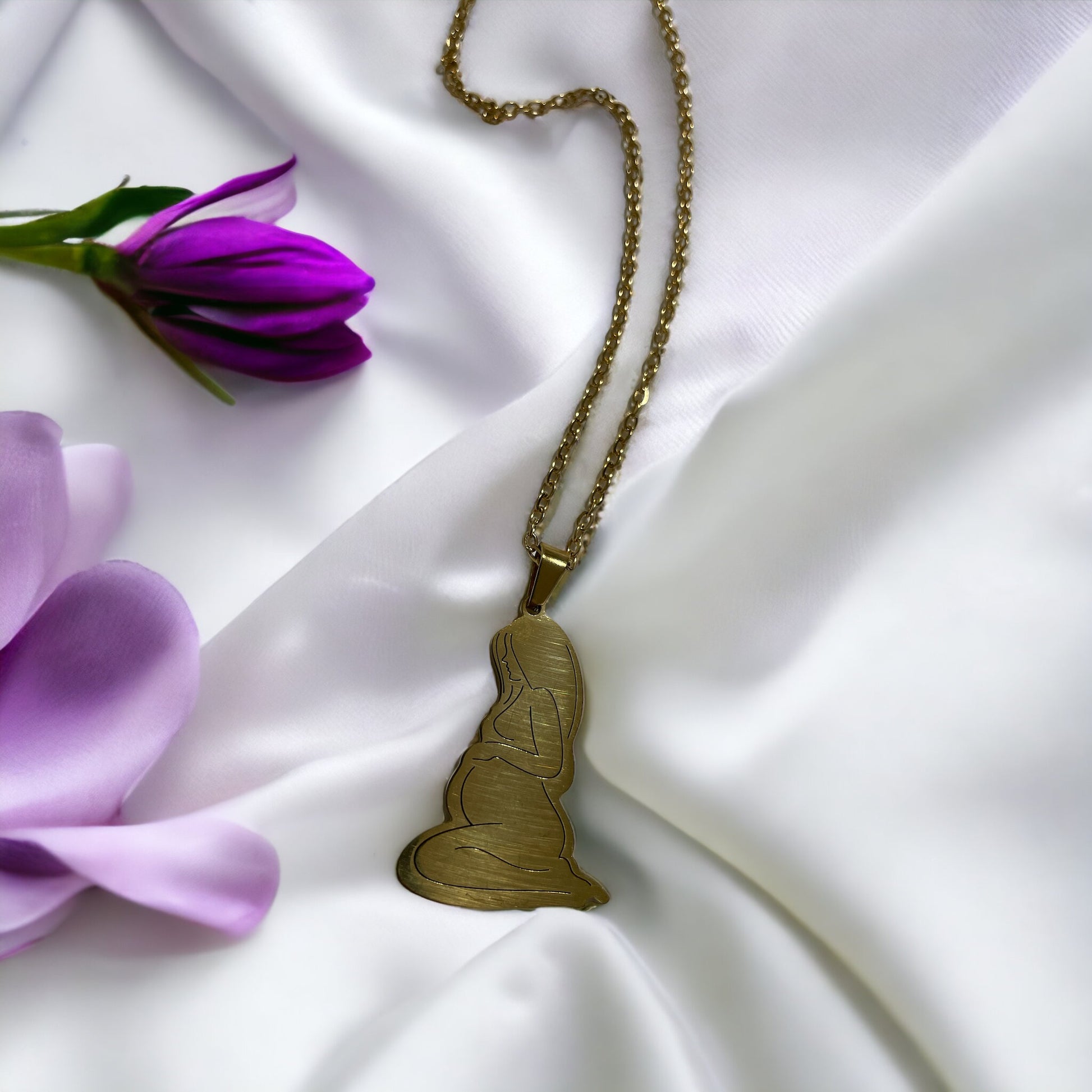 This image presents the Serene Motherhood Pendant, a delicate and polished pendant in gold plating that can also be chosen in stainless steel. The pendant features the graceful outline of a mother in a contemplative pose, cradling her child in a tender embrace, symbolizing the protective and loving bond shared. It hangs from a matching chain, showcased against a backdrop of soft, white satin that gently drapes around it, accentuating the pendant's smooth contours and reflective surface.