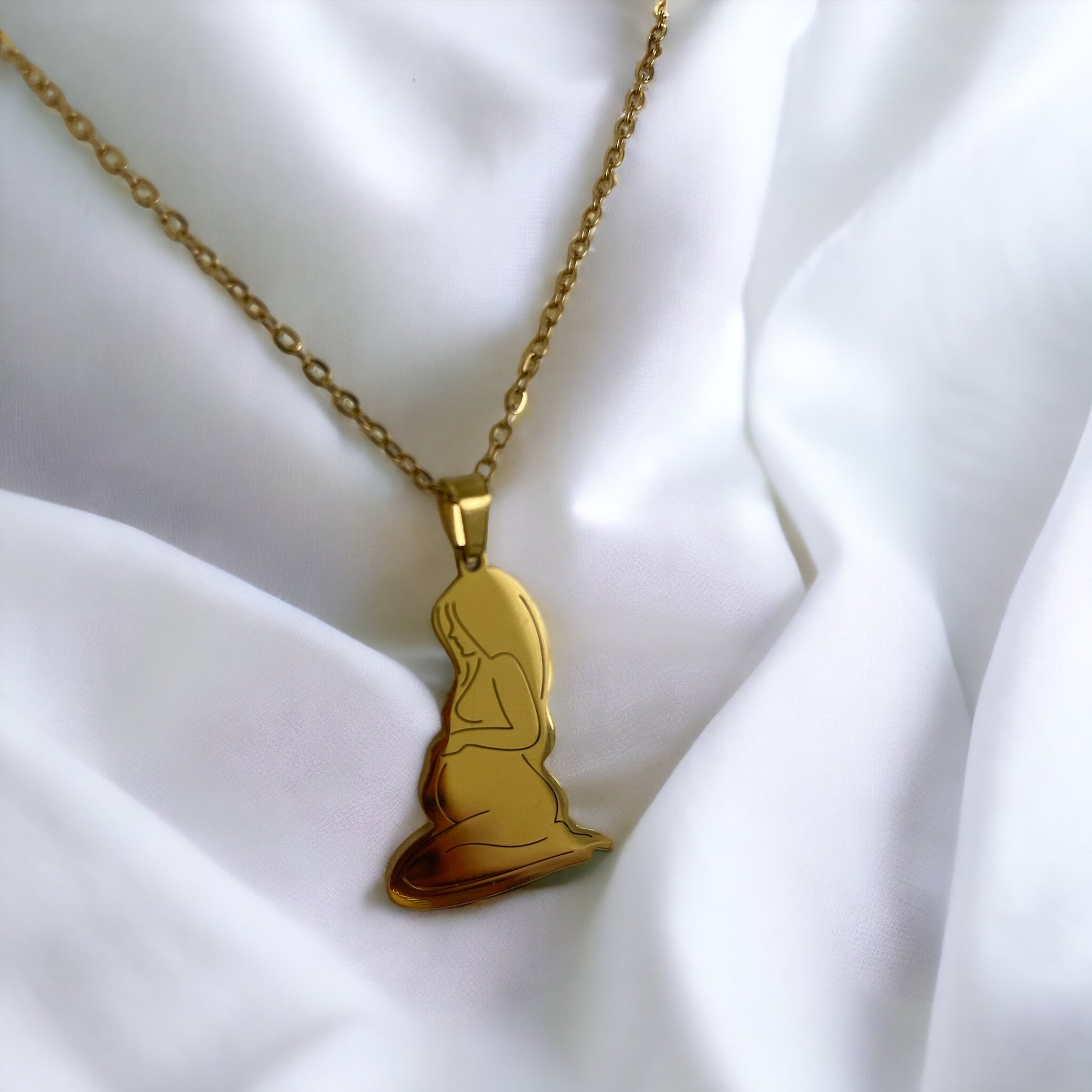 This image presents the Serene Motherhood Pendant, a delicate and polished pendant in gold plating that can also be chosen in stainless steel. The pendant features the graceful outline of a mother in a contemplative pose, cradling her child in a tender embrace, symbolizing the protective and loving bond shared. It hangs from a matching chain, showcased against a backdrop of soft, white satin that gently drapes around it, accentuating the pendant's smooth contours and reflective surface.