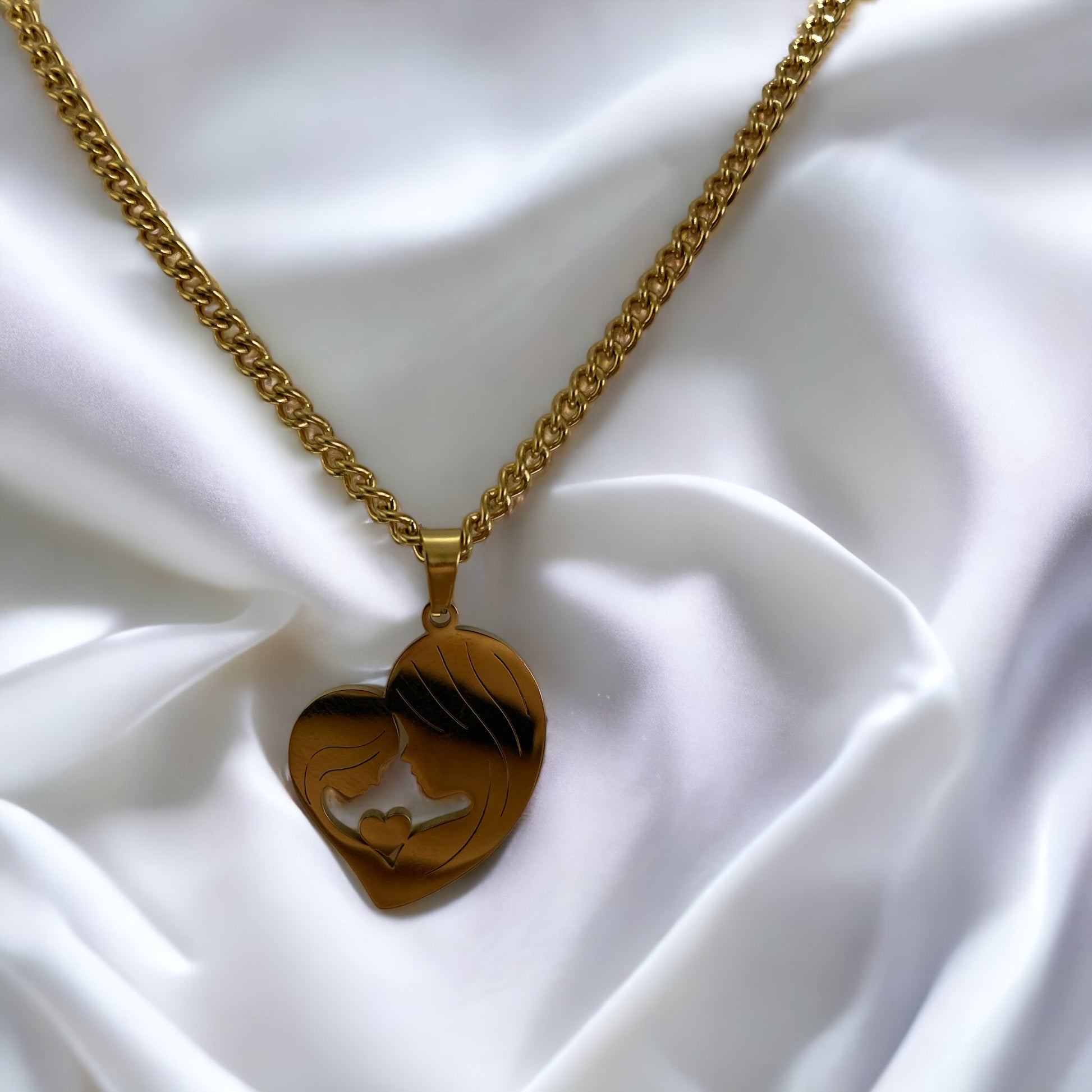 This image features the "Maternal Embrace Heart Pendant," a beautifully crafted piece with a silhouette of a mother and her child in a tender embrace, forming a heart shape. The pendant has a rich, golden hue, suggesting it is available in gold plating, with an option for stainless steel as well. It is draped over a delicate, magnolia petal, surrounded by soft, pastel flowers, enhancing the pendant's elegance and the sentiment it represents.
