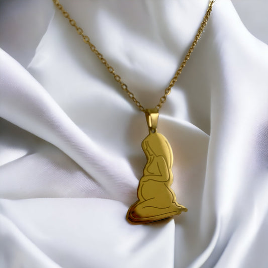 This image presents the Serene Motherhood Pendant, a delicate and polished pendant in gold plating that can also be chosen in stainless steel. The pendant features the graceful outline of a mother in a contemplative pose, cradling her child in a tender embrace, symbolizing the protective and loving bond shared. It hangs from a matching chain, showcased against a backdrop of soft, white satin that gently drapes around it, accentuating the pendant's smooth contours and reflective surface.