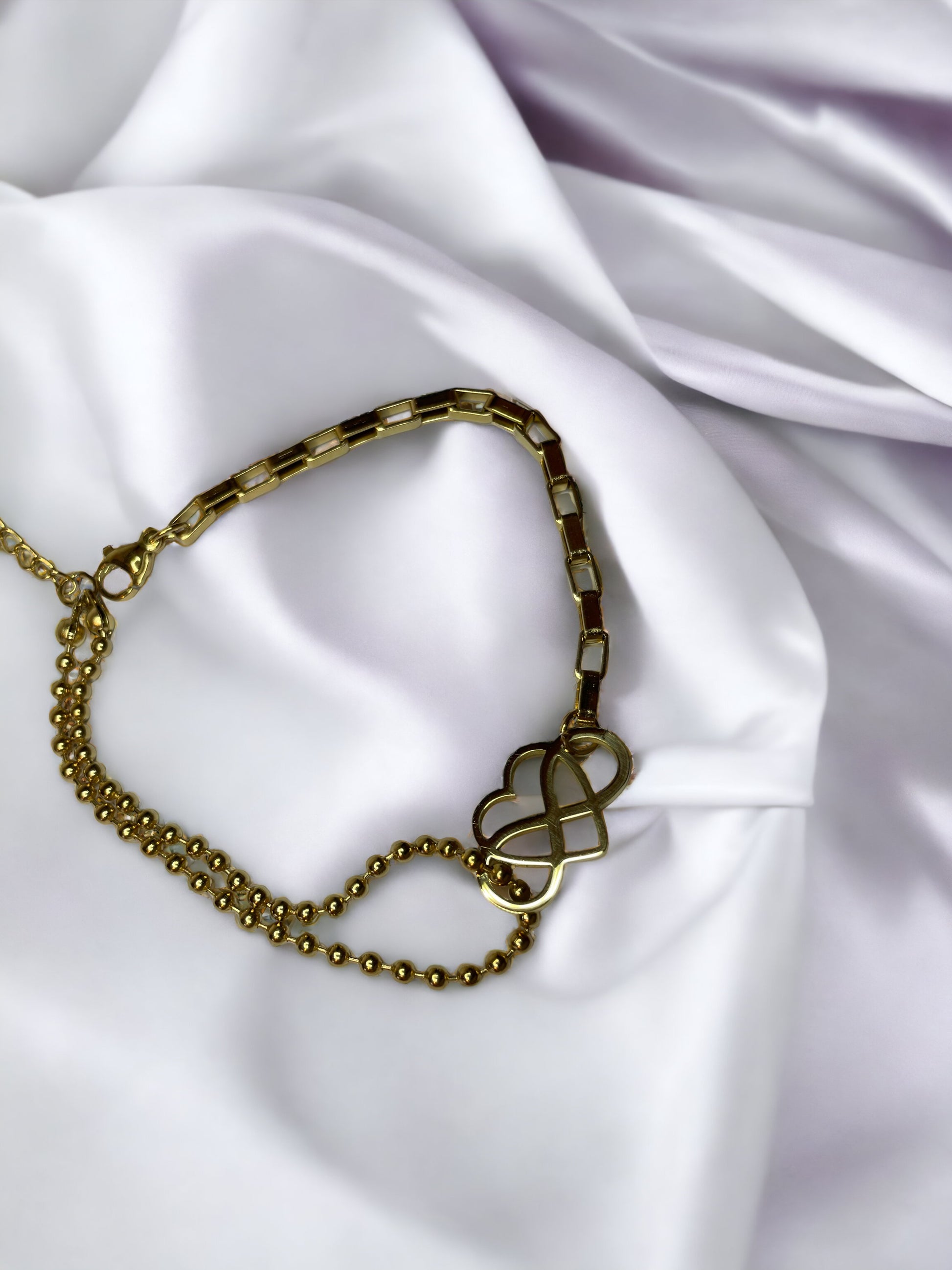 This "Eternal Embrace Bracelet" features a striking geometric heart charm that artistically intersects, symbolizing infinity and enduring love. The charm is tastefully attached to a mixed chain design composed of both a smooth, polished link chain and a rounded bead chain, adding a playful contrast to the bracelet. It is available in two finishes: a shiny stainless steel for a sleek look and a rich gold plating for a touch of warmth. 