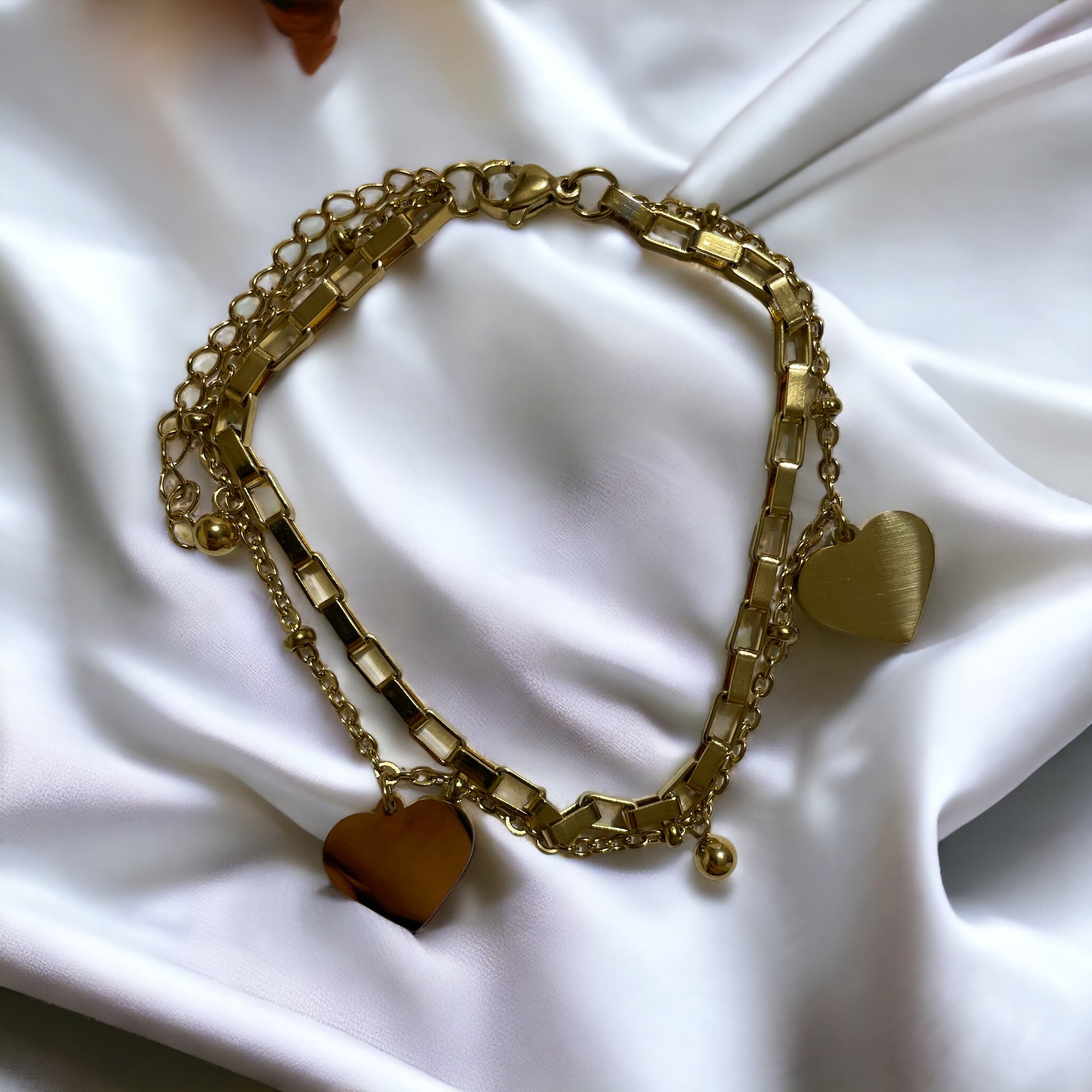 This image displays a stylish, two-layer bracelet with a polished heart charm, representing love and connection. Available in stainless steel with options for gold plating or silver finish, the bracelet combines a box chain with a beaded chain, creating a simple yet elegant accessory suitable for everyday wear or special occasions.