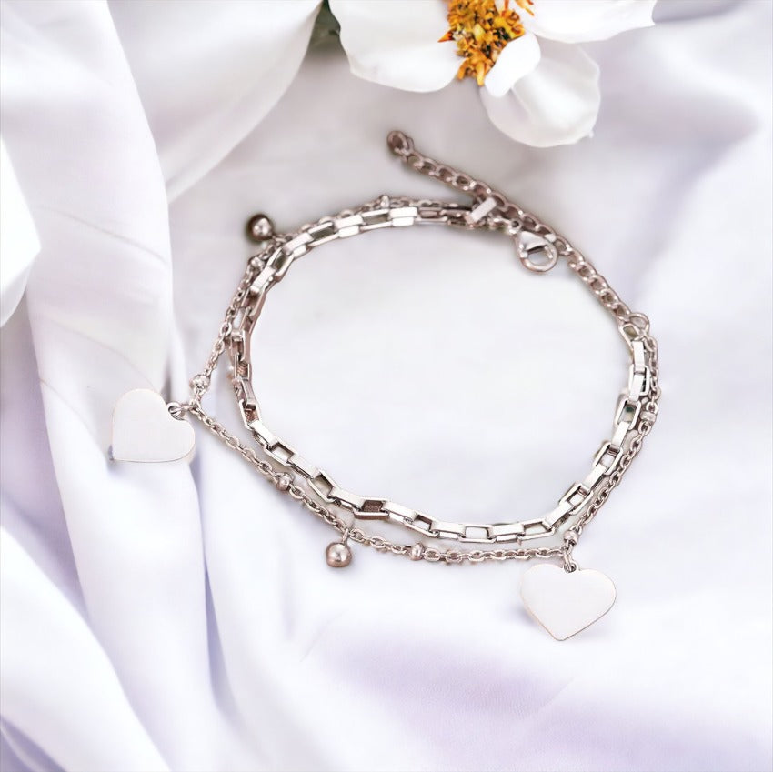 This image displays a stylish, two-layer bracelet with a polished heart charm, representing love and connection. Available in stainless steel with options for gold plating or silver finish, the bracelet combines a box chain with a beaded chain, creating a simple yet elegant accessory suitable for everyday wear or special occasions.