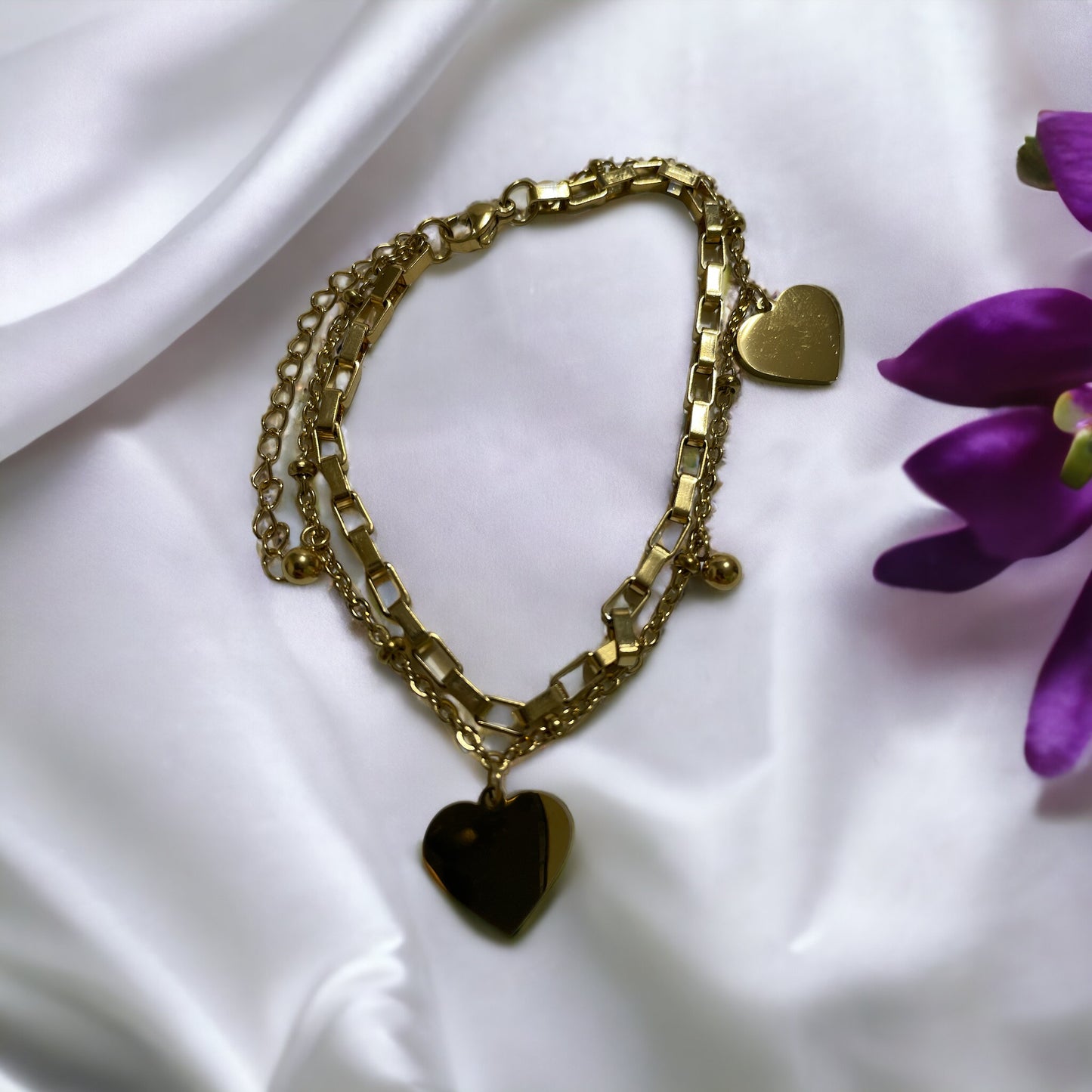 This image displays a stylish, two-layer bracelet with a polished heart charm, representing love and connection. Available in stainless steel with options for gold plating or silver finish, the bracelet combines a box chain with a beaded chain, creating a simple yet elegant accessory suitable for everyday wear or special occasions.