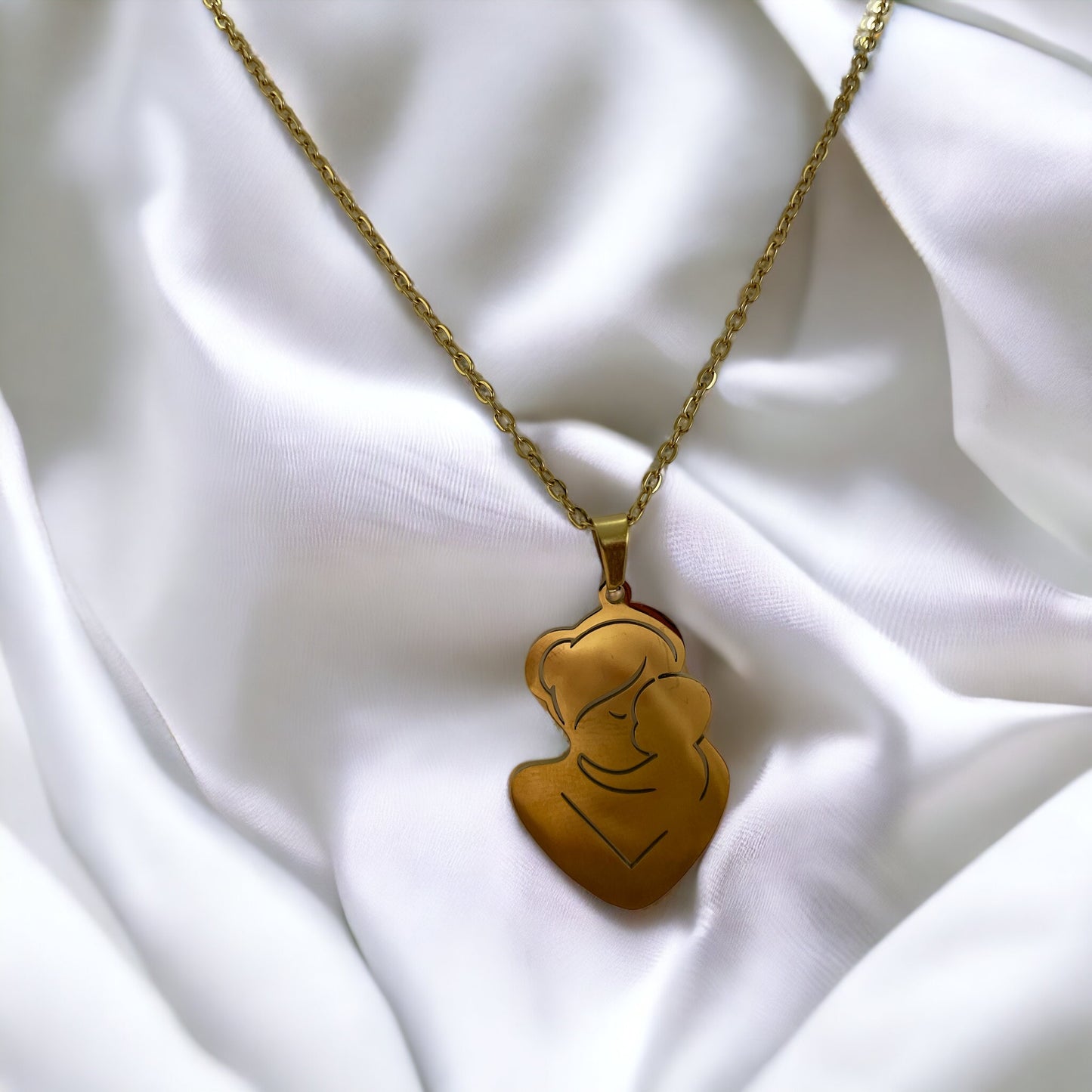The pendant is gold in color, likely gold-plated, with a silhouette design that artistically depicts a mother and child in a close, loving embrace. The smooth, shiny surface of the pendant reflects the light, giving it a warm and inviting appearance. The pendant is suspended from a delicate gold chain that complements its overall elegance. The piece exudes a sense of calm and protective love that the design intends to represent.