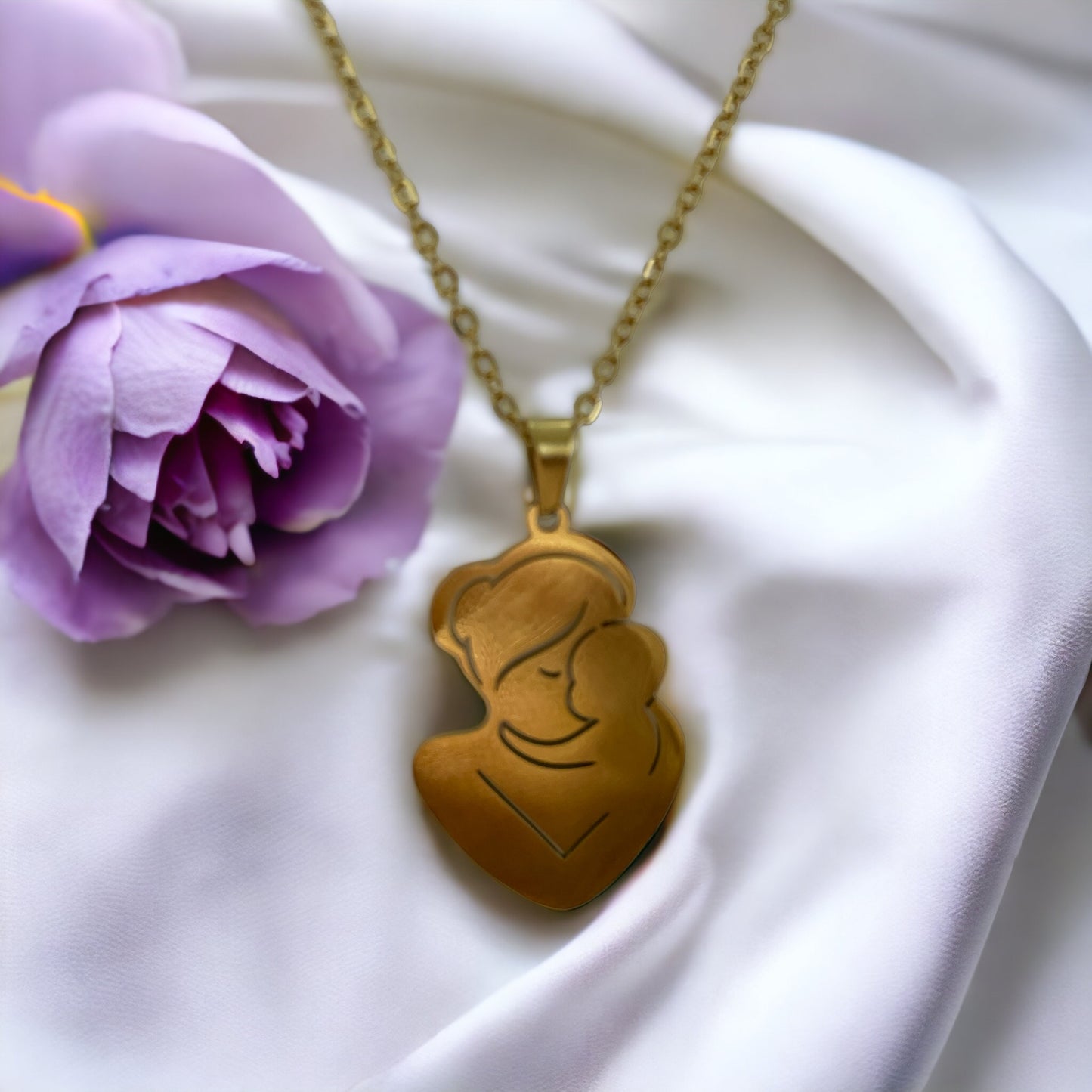 The pendant is gold in color, likely gold-plated, with a silhouette design that artistically depicts a mother and child in a close, loving embrace. The smooth, shiny surface of the pendant reflects the light, giving it a warm and inviting appearance. The pendant is suspended from a delicate gold chain that complements its overall elegance. The piece exudes a sense of calm and protective love that the design intends to represent.