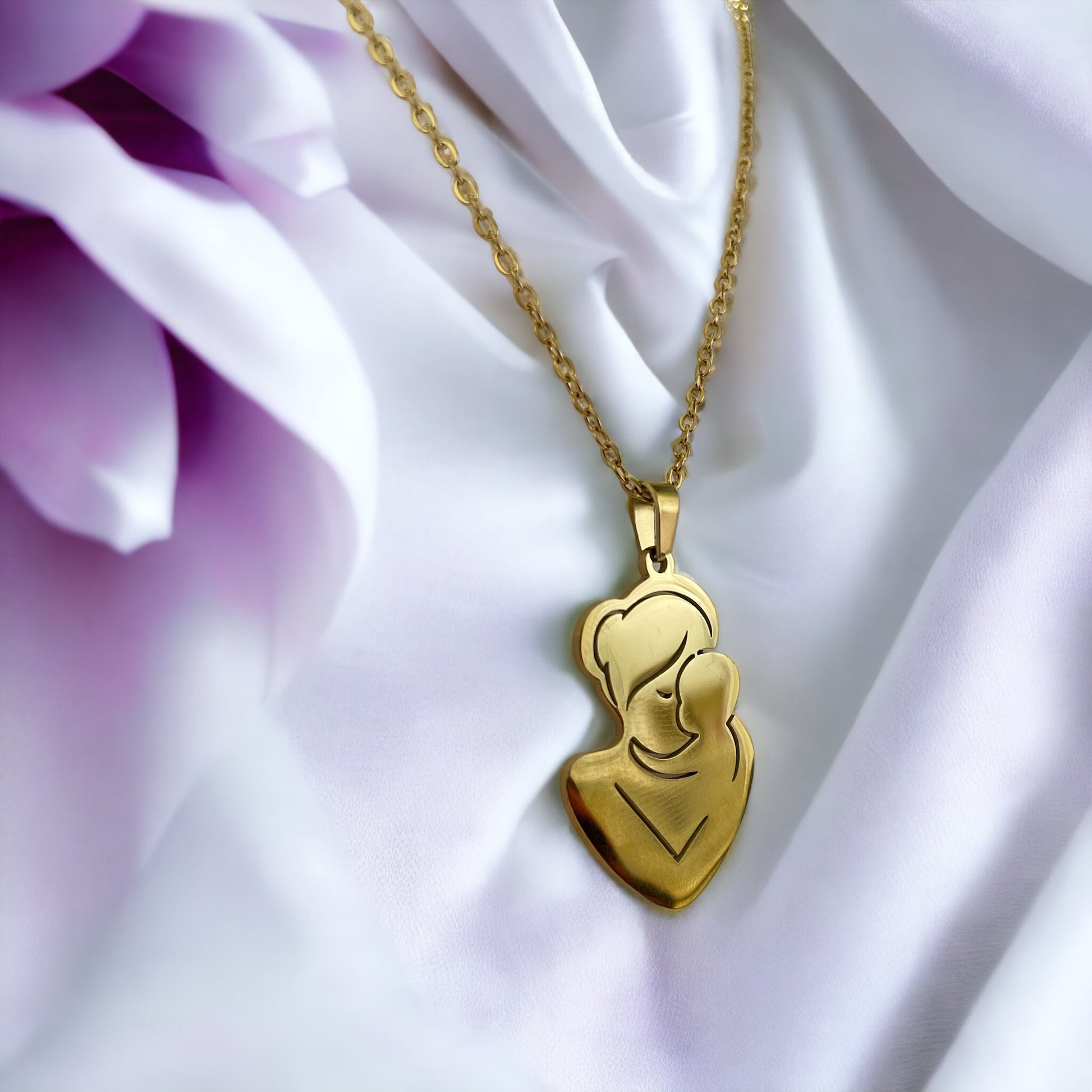 The pendant is gold in color, likely gold-plated, with a silhouette design that artistically depicts a mother and child in a close, loving embrace. The smooth, shiny surface of the pendant reflects the light, giving it a warm and inviting appearance. The pendant is suspended from a delicate gold chain that complements its overall elegance. The piece exudes a sense of calm and protective love that the design intends to represent.