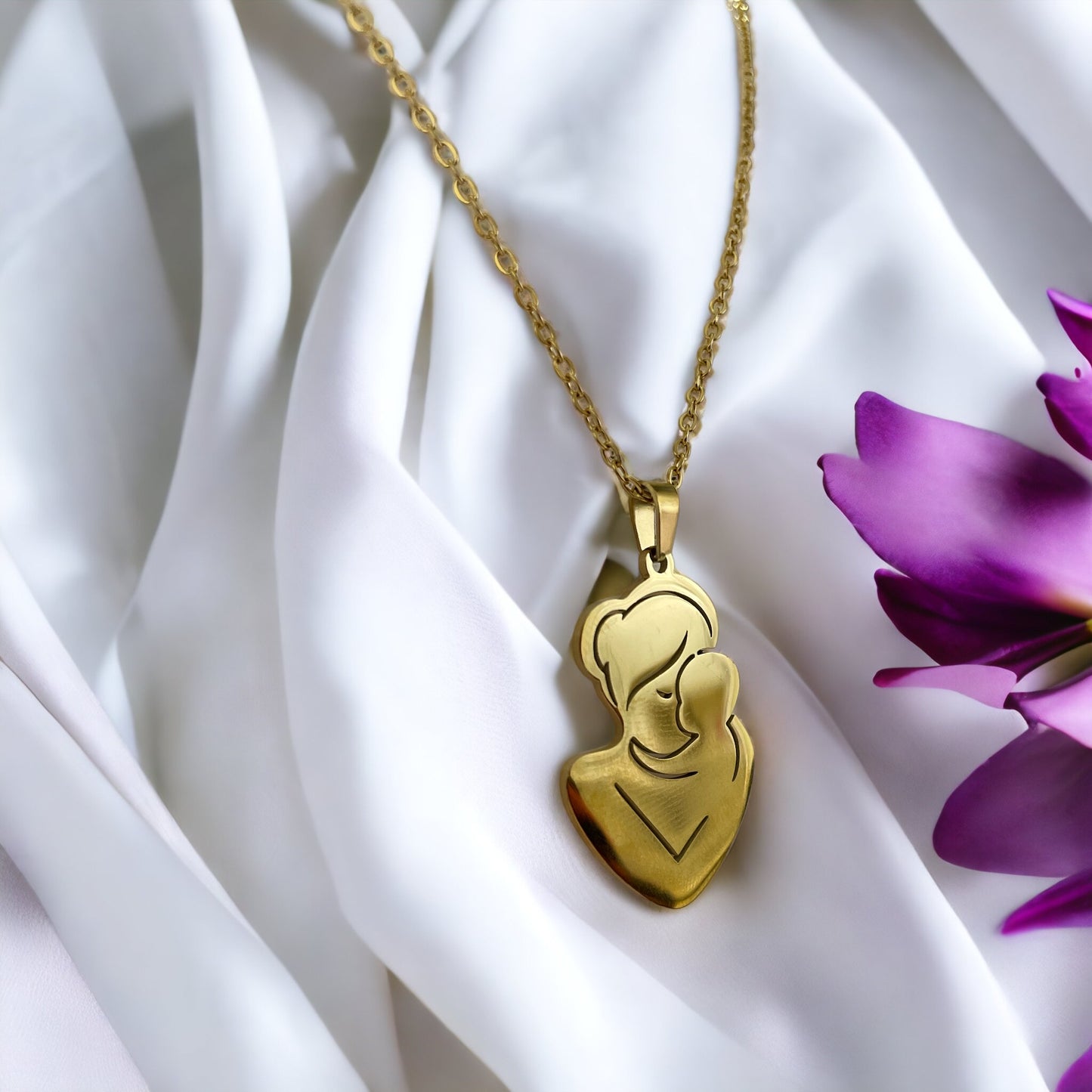 The pendant is gold in color, likely gold-plated, with a silhouette design that artistically depicts a mother and child in a close, loving embrace. The smooth, shiny surface of the pendant reflects the light, giving it a warm and inviting appearance. The pendant is suspended from a delicate gold chain that complements its overall elegance. The piece exudes a sense of calm and protective love that the design intends to represent.